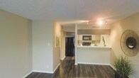 This single-floor residence offers a well-designed open layout that connects the living area to a functional kitchen, creating a cohesive and practical living environment. The dark hardwood-style flooring runs throughout the space, providing a rich contrast against the light-toned walls and white baseboards.

The kitchen is positioned at the heart of the home and features a raised breakfast bar counter that separates it from the main living area while maintaining an open feel. It is equipped with black appliances, including a full-size refrigerator, microwave, and range, complemented by white cabinetry and a decorative tile backsplash that adds a subtle design accent. A stainless sink and track lighting overhead ensure the kitchen is both practical and well-lit.

The living area is open and spacious, benefiting from a neutral color palette that enhances the sense of airiness. A textured ceiling adds dimension to the interior. A hallway leads further into the residence, offering access to additional rooms and connecting the main living space to the rest of the floor plan. The overall ambiance is clean, modern, and inviting, with thoughtful design choices that balance style and everyday functionality.