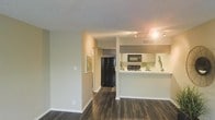 This single-floor residence offers a well-designed open layout that connects the living area to a functional kitchen, creating a cohesive and practical living environment. The dark hardwood-style flooring runs throughout the space, providing a rich contrast against the light-toned walls and white baseboards.

The kitchen is positioned at the heart of the home and features a raised breakfast bar counter that separates it from the main living area while maintaining an open feel. It is equipped with black appliances, including a full-size refrigerator, microwave, and range, complemented by white cabinetry and a decorative tile backsplash that adds a subtle design accent. A stainless sink and track lighting overhead ensure the kitchen is both practical and well-lit.

The living area is open and spacious, benefiting from a neutral color palette that enhances the sense of airiness. A textured ceiling adds dimension to the interior. A hallway leads further into the residence, offering access to additional rooms and connecting the main living space to the rest of the floor plan. The overall ambiance is clean, modern, and inviting, with thoughtful design choices that balance style and everyday functionality.