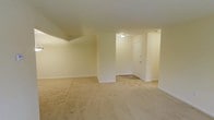This single-floor space offers an open and functional layout characterized by its neutral aesthetic and practical design. The main living area is finished with wall-to-wall beige carpeting that extends throughout, providing a soft and cohesive foundation underfoot. The walls are painted in a light, warm cream tone, contributing to a bright and airy atmosphere despite the absence of visible windows in the primary view.

A partial wall divides the main living area from an entry or transitional zone, creating a subtle sense of separation while maintaining an open flow between spaces. The entryway area features a flush-mount ceiling light fixture, illuminating the space and highlighting a standard interior door with a simple, clean design. The ceiling is smooth and unadorned, maintaining a straightforward and minimalist character throughout.

Baseboards run consistently along the perimeter of the rooms, adding a clean finishing detail. The overall layout appears well-proportioned, offering ample room for flexible furniture placement. The space presents a blank-canvas quality, making it highly adaptable for a range of interior styles and functional needs.