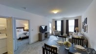 This single-floor suite offers a thoughtfully designed layout that seamlessly blends comfort and functionality. The open-plan living and dining area forms the heart of the space, bathed in natural light streaming through large windows dressed with elegant dark curtains. The dining area features a dark-wood table set for multiple guests, complete with glassware and floral accents, creating an inviting atmosphere for meals and gatherings.

Adjacent to the dining area, the living space is furnished with a plush sofa, a patterned accent chair, and a glass-top coffee table, all arranged to encourage relaxed conversation. A flat-screen television mounted on a rustic media console serves as the focal point of the entertainment area, with easy sightlines from both the living and dining zones.

The bedroom, visible through an open doorway, is a calm retreat outfitted with two beds dressed in crisp white linens and framed by warm, ambient bedside lighting. Soft carpeting runs throughout, adding a layer of warmth and comfort underfoot.

The overall aesthetic is clean and contemporary, with neutral tones across the walls and flooring complemented by coordinated furnishings. Ample natural light and an efficient, open layout make this a welcoming and versatile space suited for both short and extended stays.