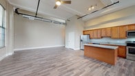 Unit 2-214 is a two-bedroom, one-bathroom residence that blends industrial-inspired architectural character with modern finishes. The open-concept living area immediately impresses with its notably high ceilings, exposed ductwork, and ceiling fan, lending an urban loft aesthetic. Large, multi-pane arched and rectangular windows flood the space with natural light while offering views of the surrounding neighborhood.

The kitchen is a functional centerpiece, featuring warm honey-toned cabinetry extending to the upper walls, granite countertops, a substantial center island with additional counter seating, and a full suite of stainless steel appliances including a refrigerator with ice maker, electric range, microwave, and dishwasher.

Both bedrooms are generously proportioned, featuring the same wood-style plank flooring found throughout the unit, neutral taupe walls, and large windows that maximize natural light. One bedroom includes a sliding door leading to an adjoining room, offering added versatility.

The bathroom is well-appointed with a granite-topped vanity, warm wood cabinetry, a full tub and shower combination with brushed nickel fixtures, and coordinating wood-style flooring, maintaining a consistent, cohesive design throughout the unit.