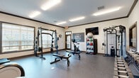 Located in Smyrna, Tennessee, this fitness center delivers a comprehensive workout facility within a single, efficiently organized space. The room features durable speckled rubber flooring throughout, providing a safe and resilient surface suited for a wide range of exercise activities. Crown molding with dark trim accents the upper walls, adding a polished architectural touch to the utilitarian space.

The facility is thoughtfully divided into distinct workout zones. A cardio section houses multiple treadmills and elliptical trainers, each positioned in front of wall-mounted flat-screen televisions, allowing members to stay entertained during their sessions. A large mirror spans the wall in this area, enhancing the sense of openness. Adjacent to this is a strength training zone featuring a full power rack with weight plates and adjustable benches, a cable machine, and a dedicated dumbbell rack with a full range of free weights.

A separate area provides space for functional training, equipped with a stationary bike, medicine balls, kettlebells, and a stability ball neatly organized on a multi-tier rack. A motivational "Stay Humble, Hustle Hard" wall graphic adds an energetic atmosphere. Natural light filters in through multiple windows fitted with horizontal blinds, while flush fluorescent ceiling fixtures ensure consistent illumination throughout. A glass-paneled entry door completes the clean, welcoming design.
