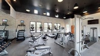 This single-level fitness center offers a thoughtfully designed workout environment that balances functionality with modern aesthetics. The space is defined by its striking contrast of a dark-painted ceiling against white and exposed brick walls, creating a bold, contemporary atmosphere. Pendant lighting fixtures hang from the ceiling alongside recessed lights, providing well-distributed illumination throughout the facility.

The cardio zone lines the brick-accented wall and features a row of treadmills, ellipticals, and upright bikes, all equipped with individual screens. Multiple windows along this wall allow natural light to fill the space while offering outdoor views. A large mirrored wall on the opposite side visually expands the floor plan and reflects the full range of equipment.

The strength training area is generously outfitted with Life Fitness cable machines, a full dumbbell rack, adjustable benches, and a variety of weight machines targeting multiple muscle groups. Glass doors on one side of the facility provide additional natural light and direct access to an outdoor area. A bank of wooden lockers offers convenient storage, and wall-mounted flat-screen TVs are positioned throughout for entertainment during workouts. Durable rubber and carpet-tile flooring complete the practical, gym-grade interior.