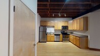 This single-floor urban loft showcases a striking industrial aesthetic defined by exposed brick and concrete block walls, rich dark-stained wood beam ceilings, and light hardwood flooring throughout. The open living area is a standout space, featuring dramatic exposed ductwork, a ceiling fan, and a sliding glass door that leads to a private balcony overlooking the surrounding cityscape, filling the room with natural light. A carpeted entry area transitions seamlessly into the main living space, adding a layer of texture to the overall design. The kitchen is well-equipped with an L-shaped layout offering ample storage through warm-toned wood cabinetry, granite-style countertops, and a full suite of stainless steel appliances including a refrigerator, electric range, dishwasher, and microwave. Track lighting mounted on the exposed beam ceiling provides targeted illumination throughout the kitchen. The bathroom features a single-vanity with wood cabinetry, a large mirror, a combined tub and shower, and a generously sized open shelving unit with wire racks, offering practical storage solutions. The overall ambiance of this loft is distinctly urban and industrial, blending raw architectural elements with functional modern finishes.