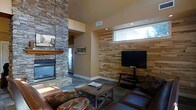 This single-floor space showcases a thoughtfully designed living area that blends rustic and contemporary elements. The room is anchored by a commanding floor-to-ceiling stacked stone fireplace with a warm wood mantel, serving as a dramatic focal point that adds texture and character to the space. Adjacent to the fireplace, a reclaimed wood plank accent wall provides a natural, earthy backdrop for the mounted flat-screen television and media console, creating a cohesive entertainment area.

The living space is furnished with rich brown leather seating and a rustic wooden coffee table, reinforcing the warm, cabin-inspired aesthetic. A neutral color palette on the walls complements the natural materials throughout, while recessed lighting and a horizontal clerestory window allow natural light to filter into the space, enhancing its openness despite the cozy atmosphere.

The flooring features grey tile that grounds the room and adds a modern contrast to the warmer wood and stone elements. Overall, the space strikes a balance between rugged mountain-style charm and comfortable, modern living, making it well-suited as a retreat or relaxation-focused residence.
