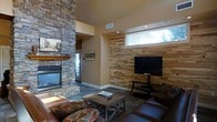 This single-floor space showcases a thoughtfully designed living area that blends rustic and contemporary elements. The room is anchored by a commanding floor-to-ceiling stacked stone fireplace with a warm wood mantel, serving as a dramatic focal point that adds texture and character to the space. Adjacent to the fireplace, a reclaimed wood plank accent wall provides a natural, earthy backdrop for the mounted flat-screen television and media console, creating a cohesive entertainment area.

The living space is furnished with rich brown leather seating and a rustic wooden coffee table, reinforcing the warm, cabin-inspired aesthetic. A neutral color palette on the walls complements the natural materials throughout, while recessed lighting and a horizontal clerestory window allow natural light to filter into the space, enhancing its openness despite the cozy atmosphere.

The flooring features grey tile that grounds the room and adds a modern contrast to the warmer wood and stone elements. Overall, the space strikes a balance between rugged mountain-style charm and comfortable, modern living, making it well-suited as a retreat or relaxation-focused residence.