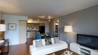 This single-floor space showcases an open-concept design that seamlessly integrates the living, dining, and kitchen areas into one cohesive environment. The living area is anchored by a contemporary white sofa adorned with decorative throw pillows, paired with a glass coffee table and a tall floor lamp that provides warm ambient lighting. Light grey walls complement the bright white ceiling, giving the space an airy, clean aesthetic.

The dining area sits adjacent to the living space, featuring a wooden table surrounded by blue upholstered chairs, creating a casual yet inviting setting for meals. A fully equipped kitchen lines the background, offering warm wood cabinetry with open upper shelving, a dark refrigerator, and a functional countertop workspace. A mirrored backsplash above the sink adds depth and reflects light throughout the area.

Track lighting installed along the ceiling provides targeted illumination across the open-plan layout, enhancing visibility and ambiance. A closet corridor with shelving is visible toward the back, indicating practical storage solutions. The warm hardwood flooring throughout ties all zones together, reinforcing the unit's cohesive and modern character.