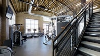 Located at 4101 South Navajo Street in Englewood, Colorado, this two-story fitness facility combines industrial aesthetics with functional design. The space is characterized by its exposed ceiling joists, visible ductwork, and warm brick walls, creating a raw yet polished atmosphere that speaks to the building's character.

The main gym floor is generously proportioned, offering ample room for a comprehensive array of exercise equipment. Cardio machines — including treadmills, ellipticals, and stair climbers — are positioned near large windows that invite natural light into the space, creating an energizing workout environment. Strength training is well-represented with multi-station cable machines, free weights, and benches arranged across the floor.

A metal-railed staircase with mesh detailing connects the two levels, reinforcing the industrial design language throughout. The upper level overlooks the main floor, providing an open, connected feel between the two spaces. Polished concrete flooring throughout adds to the utilitarian yet refined character of the facility.

The combination of high ceilings, natural light from oversized windows, exposed structural elements, and modern fitness equipment makes this a well-appointed and visually distinctive training environment suited to a wide range of fitness activities.