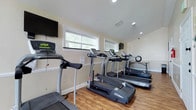 This fitness center is a comprehensively outfitted exercise facility designed to support a full range of workout routines. The space is finished with warm hardwood-style flooring throughout, neutral beige walls, white wainscoting, and recessed lighting that creates a clean, well-lit environment. Multiple windows allow natural light to filter in, contributing to an open and energizing atmosphere.

The cardio area is stocked with several treadmills — including Bowflex and Vision models — along with a stationary bike and an elliptical trainer. Wall-mounted flat-screen televisions are positioned throughout the space, allowing users to stay entertained during cardio sessions. A utility table with a wall clock provides a convenient spot for personal items.

The strength training section features a multi-function cable machine with a weight bench, a barbell rack, and a dedicated free weights area equipped with kettlebells and dumbbells on a rack. Resistance bands and suspension training straps are mounted to the wall, offering additional functional training options. Large mirrors line one wall of the strength area, maximizing the sense of space and aiding in form correction.

Protective rubber mats are placed beneath heavy equipment, and the layout allows ample room to move freely between stations. An emergency exit door provides convenient access, rounding out this practical and well-organized fitness facility.