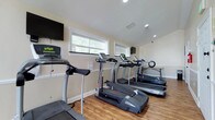 This fitness center is a comprehensively outfitted exercise facility designed to support a full range of workout routines. The space is finished with warm hardwood-style flooring throughout, neutral beige walls, white wainscoting, and recessed lighting that creates a clean, well-lit environment. Multiple windows allow natural light to filter in, contributing to an open and energizing atmosphere.

The cardio area is stocked with several treadmills — including Bowflex and Vision models — along with a stationary bike and an elliptical trainer. Wall-mounted flat-screen televisions are positioned throughout the space, allowing users to stay entertained during cardio sessions. A utility table with a wall clock provides a convenient spot for personal items.

The strength training section features a multi-function cable machine with a weight bench, a barbell rack, and a dedicated free weights area equipped with kettlebells and dumbbells on a rack. Resistance bands and suspension training straps are mounted to the wall, offering additional functional training options. Large mirrors line one wall of the strength area, maximizing the sense of space and aiding in form correction.

Protective rubber mats are placed beneath heavy equipment, and the layout allows ample room to move freely between stations. An emergency exit door provides convenient access, rounding out this practical and well-organized fitness facility.