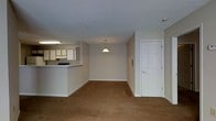 This single-floor space showcases an open-concept design that connects the kitchen and living area into one cohesive layout. The living area is finished with warm-toned carpet flooring and neutral-painted walls, creating a clean, understated aesthetic. A textured popcorn ceiling runs throughout, and a pendant light fixture is centrally positioned, providing focused illumination to the main living space.

The kitchen is situated in the corner of the unit, separated from the living area by a raised breakfast bar counter that offers a natural division between the two zones while maintaining the open feel. White cabinetry lines the kitchen walls, complemented by under-cabinet lighting that brightens the workspace. A built-in stovetop is visible along the counter, and the overhead fluorescent light fixture provides additional task lighting.

A white six-panel door near the corner of the living area leads to an adjacent room or exterior entry point. The overall ambiance of the space is practical and straightforward, with a muted, neutral color palette throughout that offers a versatile and move-in-ready environment.