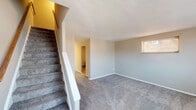 This two-floor residence offers a practical and straightforward layout spread across two levels, connected by a carpeted staircase with a white-painted banister and a warm wood-toned handrail. The main level welcomes you through a front entry door with a glass panel, opening into a spacious living area finished with neutral gray carpet and light-toned walls. A window allows natural light to filter into the room, and an adjacent hallway leads further into the home. The kitchen is compact yet functional, equipped with a gas range and a full-size refrigerator, both in white, along with wood-look vinyl flooring and a window overlooking the exterior. A full bathroom on this level features a tub-shower combination, a toilet, and wood-look flooring, with a small window providing ventilation and light. Upstairs, the bedroom showcases a distinctive angled ceiling that follows the roofline, adding architectural character to the space. The room is finished with soft beige carpet and neutral walls, creating a quiet and comfortable retreat. Throughout the home, the design emphasizes clean, simple finishes with a neutral color palette, offering a move-in ready environment adaptable to a variety of personal styles.