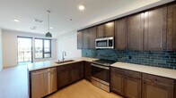 This single-floor residence showcases a thoughtfully designed interior with modern finishes throughout. The kitchen serves as a centerpiece of the space, featuring rich dark wood cabinetry, crisp white quartz countertops, and a striking teal subway tile backsplash that adds a bold pop of color. A suite of stainless steel appliances — including a gas range, built-in microwave, refrigerator, and dishwasher — provides full functionality. A dark-paneled kitchen island with a built-in sink and pendant lighting overhead adds both style and additional workspace. Light wood-tone flooring extends throughout the open-plan kitchen and living areas, lending warmth and continuity to the space. Natural light filters in through large windows that frame outdoor views of greenery, enhancing the bright, airy ambiance. The bedroom is finished with plush carpeting and features a floor-to-ceiling window with balcony access, offering scenic outdoor views. The bathroom is finished with large-format gray floor tiles, a sleek sliding-glass walk-in shower adorned with vertical white subway tiles, a white quartz vanity countertop, and dark wood cabinetry — creating a clean, contemporary aesthetic. Overall, the space combines modern design elements with practical finishes for a polished, comfortable living environment.