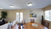 The Recreation Room at 1394 Oro Vista in San Diego is a welcoming, single-floor community space designed to accommodate gatherings and social activities. The main area is generously sized, featuring warm hardwood-style flooring and neutral-toned walls that create a bright, inviting atmosphere. Multiple round dining tables with dark wood chairs are arranged throughout the room, offering ample seating for residents and guests. Large windows along the walls allow abundant natural light to fill the space, while decorative artwork — including a striking Eiffel Tower print — adds a touch of character. Flush-mount ceiling light fixtures with a textured ceiling provide consistent overhead lighting throughout. A comfortable lounge area is also present, furnished with a dark leather sofa, a plush area rug, and accent pieces including a decorative mirror, offering a more relaxed setting for socializing. French doors lead to an outdoor balcony area, further enhancing the sense of openness. A functional kitchenette supports the space, equipped with a full-size refrigerator, a sink with cabinetry, and granite-style countertops — ideal for hosting events. Overall, this recreation room provides a practical and comfortable environment for community gatherings.