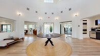 This expansive community clubhouse delivers a well-rounded amenity experience across a generously sized single floor. The dramatic main lobby sets the tone immediately, showcasing soaring ceilings, abundant natural light filtering through high clerestory windows, and light wood flooring that flows throughout. A statement round table anchored by a woven jute rug serves as the centerpiece of this welcoming entry space.

From the lobby, residents can access a range of thoughtfully designed areas. The fitness center is well-equipped with a full lineup of cardio machines, strength-training equipment, and free weights, complemented by floor-to-ceiling mirrors, ceiling fans, and wall-mounted TVs. A dedicated conference room with glass door entry offers a polished setting for meetings, furnished with a large work table, ergonomic seating, and a mounted display screen.

The game room provides a lively social hub, complete with a pool table, shuffleboard, a wet bar area with bar seating, and views of the surrounding tree canopy through multiple windows. A stylish coffee bar station with cabinetry, a professional espresso machine, and open shelving adds a convenient hospitality touch.

Outside, the property features a beautifully landscaped courtyard with flagstone steps, natural stone exterior walls, a covered cabana with lounge seating, and mature oak trees — creating a true resort-style retreat.