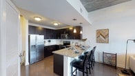 This single-floor space showcases a thoughtfully designed open-concept layout that seamlessly blends style and functionality. The kitchen is a standout feature, outfitted with rich dark espresso cabinetry that extends to the ceiling, providing generous storage throughout. Gleaming granite countertops offer ample prep space and carry a sophisticated aesthetic, while a large peninsula with seating for multiple bar-height chairs serves as both a social hub and a practical dining area. Stainless steel appliances, including a French-door refrigerator with ice and water dispenser, built-in microwave, and range, complete the contemporary culinary space. Pendant lighting suspended above the peninsula adds warm, focused illumination, complementing the flush-mount ceiling fixtures. The kitchen flows naturally into the living area, reinforcing the open feel of the floor plan. Hardwood flooring runs throughout, adding warmth and continuity to the space. A white subway tile backsplash provides a clean contrast against the dark cabinetry, while an exposed concrete ceiling detail adds an industrial touch. The overall ambiance is modern and polished, balancing dark tones with bright, neutral walls and thoughtful lighting to create an inviting and stylish environment.