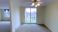 This single-floor space is thoughtfully designed with comfort and functionality in mind. The main living area features soft, neutral-toned carpeting underfoot and smooth, light-colored walls that create a bright, open atmosphere. A ceiling fan with an integrated light fixture is centrally positioned, providing both air circulation and ambient lighting throughout the room.

A prominent sliding glass door serves as a key architectural feature, flooding the interior with generous natural light and offering direct access to a private balcony. The balcony is enclosed by a wrought iron railing and provides views of a green lawn and surrounding trees, adding a pleasant outdoor connection to the living space. The exterior brick detailing visible through the glass adds a sense of architectural character.

A baseboard heating unit runs along the exterior wall, ensuring efficient climate control throughout the colder months. The overall ambiance of the space is calm and understated, with its neutral palette and natural light making it a versatile and welcoming environment. The seamless indoor-outdoor connection through the balcony access is a notable highlight of this space.