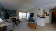 This single-floor residence offers a comfortable and functional layout centered around an open-concept living space. The living room is a welcoming area highlighted by a classic white-mantled fireplace, which serves as a focal point along a teal accent wall adorned with decorative framing. Large windows and a set of French doors flood the room with natural light and provide views of the surrounding outdoor greenery, creating a bright and airy ambiance. The living area is finished with neutral carpeting that adds warmth underfoot, and features a textured ceiling that adds subtle architectural character. A low half-wall partition seamlessly connects the living room to the kitchen, which is equipped with light wood cabinetry, a countertop range hood, and ample counter space — making it ideal for both everyday cooking and casual entertaining. The open sightlines between the kitchen and living room enhance the sense of space throughout. Overall, the residence presents a cohesive, well-lit interior with a practical flow, combining cozy residential touches with functional design elements suited for comfortable everyday living.
