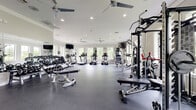Located at 8940 N Congress Ave in Kansas City, MO, this fitness center offers a spacious, single-floor layout designed to accommodate a comprehensive range of workout activities. The open floor plan provides generous room to move between equipment zones without feeling crowded, all finished with durable rubber flooring suited for high-impact exercise.

The cardio section is lined with treadmills, ellipticals, and stair climbers positioned along a wall of large windows, which flood the space with abundant natural light and offer views of the surrounding greenery. Full-length mirrors are strategically placed throughout, enhancing the sense of openness while allowing users to monitor their form.

The strength training area is well-appointed with a multi-station cable machine, adjustable weight benches, and a fully stocked dumbbell rack featuring a wide range of weights. A dedicated functional fitness zone houses stability balls, resistance bands, and additional accessories for varied training routines. A water fountain is conveniently accessible within the space.

Overhead, multiple ceiling fans complement the recessed lighting, maintaining airflow and comfort throughout the facility. Wall-mounted flat-screen televisions are positioned at multiple points for entertainment during workouts. The clean, neutral color palette and bright, airy atmosphere create an inviting and motivating environment for all fitness levels.