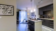 This single-floor residence offers a thoughtfully designed open-concept layout that seamlessly connects the kitchen, dining, and living areas into one cohesive living space. The kitchen is outfitted with dark cabinetry, light stone countertops, and a peninsula with bar seating illuminated by sleek pendant lighting — ideal for casual dining or entertaining. Track lighting above provides additional brightness to the culinary space. The living area is anchored by smooth hardwood-style flooring and features a ceiling fan, adding both comfort and function. A large area rug defines the seating zone, which is bathed in natural light streaming through floor-to-ceiling sliding glass doors dressed with sheer vertical blinds. These doors open to an outdoor space, blending interior and exterior living effortlessly. The walls are finished in a neutral palette, complemented by contemporary décor accents including framed wall art. The overall ambiance is modern and airy, with clean lines, layered lighting, and an open flow that makes the space feel welcoming and versatile.