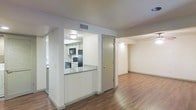 This single-floor space is designed with an open, functional layout that maximizes the sense of space throughout. The living area features warm hardwood flooring that extends throughout, complementing the clean white walls and creating a bright, welcoming atmosphere. A semi-open kitchen is integrated into the layout via a pass-through counter finished with granite-style countertops, offering both a visual connection to the living space and practical utility. The kitchen is equipped with black appliances, including a refrigerator, microwave, and electric range, alongside white cabinetry that keeps the space feeling light and organized. A door adjacent to the kitchen provides access to an additional room or utility area, contributing to the overall flow of the floor plan. The living area extends generously beyond the kitchen partition, offering ample room for furnishing and everyday use. Recessed and flush-mount lighting fixtures are positioned throughout, ensuring consistent illumination across the space. The overall ambiance is clean, neutral, and move-in ready, making it a versatile and practical living environment.