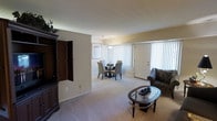 This single-floor residence presents an open-concept layout that seamlessly connects the living and dining areas, creating a spacious and cohesive environment. The interior is finished with wall-to-wall plush carpeting in a warm neutral tone, complementing the soft beige walls throughout. The living area is anchored by a large dark wood entertainment unit with cabinet doors, providing generous storage and display space. A glass-topped oval coffee table adds a refined touch to the seating area, which also features an upholstered accent chair. The adjoining dining area is defined by a circular dining table surrounded by upholstered chairs, positioned beneath an elegant pendant light fixture. A framed piece of wall art adds a decorative accent to this zone. Large windows dressed with vertical blinds allow natural light to filter generously into the space, contributing to its bright and airy ambiance. A full-panel white door provides access to the exterior, further enhancing the flow of light. The overall design maintains a clean, understated aesthetic with consistent finishes that create a welcoming and functional living environment throughout.