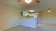 This single-floor residence offers a practical and functional open-concept design that connects the living area and kitchen in a cohesive, flowing layout. The living area is finished with neutral-toned carpet and features a ceiling fan with a light fixture, providing both comfort and overhead illumination. A peninsula-style breakfast bar serves as the natural dividing point between the living space and the kitchen, offering casual seating and additional counter space. The kitchen is outfitted with ample white cabinetry that extends to the upper walls, providing generous storage, along with a light-toned countertop and white appliances including a refrigerator. A fluorescent overhead light fixture ensures the kitchen is well-lit for meal preparation. The walls throughout are painted in a soft, neutral tone that contributes to the bright and airy ambiance of the space. The open arrangement between the kitchen and living area promotes ease of movement and a sense of spaciousness, making the layout well-suited for both daily living and casual entertaining. The clean, understated finishes throughout give the space a move-in-ready quality.