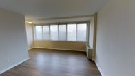 This single-floor space offers a clean and functional layout designed for comfortable living. The main living area is a standout feature, characterized by a wide bank of windows fitted with horizontal blinds that allow for generous amounts of natural light to fill the room. The windows span a significant portion of the wall, providing an airy, open feel to the interior. The flooring throughout is a warm hardwood-style laminate in a medium-toned finish, adding a sense of warmth and continuity to the space. The walls are painted in a soft, neutral yellow-white tone that complements the light wood flooring and contributes to the overall bright ambiance. A wall-mounted HVAC unit is positioned beneath the windows, offering climate control directly within the living area. The ceiling is smooth and white, with recessed flush lighting providing additional illumination. White baseboards and clean wall finishes complete the polished, well-maintained appearance of the space. Overall, the unit presents a practical and inviting environment, well-suited for those seeking a bright, low-maintenance living space with a simple, modern aesthetic.