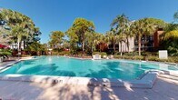 This expansive outdoor amenity area centers around a generously sized swimming pool with clear turquoise water, framed by a broad concrete pool deck. The pool features a distinctive angular design with gradual depth markings, accommodating a range of swimmers. A row of chaise lounges with red cushions lines the deck, accompanied by shaded seating areas with red umbrellas, providing ample space for sunbathing and relaxation.

Adjacent to the main pool, a separate spa offers a more intimate setting, with nearby lounge seating arranged for quiet enjoyment. A dedicated support building with a metal roof sits at the edge of the deck, providing convenient access to amenities.

The surrounding landscape is a standout feature of this outdoor space. Tall Florida pines, swaying palm trees, banana plants, and vibrant tropical foliage — including richly colored crotons and ornamental shrubs — create a lush, resort-like atmosphere. Mature trees provide natural shade and privacy throughout the area. The layered plantings and verdant greenery lend a secluded, tropical character to the entire setting, making the outdoor area feel like a private retreat under the broad Florida sky.