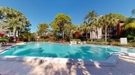 This expansive outdoor amenity area centers around a generously sized swimming pool with clear turquoise water, framed by a broad concrete pool deck. The pool features a distinctive angular design with gradual depth markings, accommodating a range of swimmers. A row of chaise lounges with red cushions lines the deck, accompanied by shaded seating areas with red umbrellas, providing ample space for sunbathing and relaxation.

Adjacent to the main pool, a separate spa offers a more intimate setting, with nearby lounge seating arranged for quiet enjoyment. A dedicated support building with a metal roof sits at the edge of the deck, providing convenient access to amenities.

The surrounding landscape is a standout feature of this outdoor space. Tall Florida pines, swaying palm trees, banana plants, and vibrant tropical foliage — including richly colored crotons and ornamental shrubs — create a lush, resort-like atmosphere. Mature trees provide natural shade and privacy throughout the area. The layered plantings and verdant greenery lend a secluded, tropical character to the entire setting, making the outdoor area feel like a private retreat under the broad Florida sky.