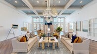 This thoughtfully designed single-story space functions as a sophisticated communal facility, blending coastal elegance with modern amenities across several distinct areas. The centerpiece is a grand, open lounge featuring soaring coffered ceilings painted in a soft blue-gray tone, anchored by a dramatic cluster of crystal chandeliers. Floor-to-ceiling windows and multiple sets of French doors flood the room with natural light, while cream sofas accented with navy and mustard pillows create inviting seating arrangements around a marble-topped coffee table. A striking shiplap fireplace wall with a veined stone surround and mounted television serves as a focal point on one end, with French doors leading to an adjacent game room housing a full-sized billiards table, warm wood pendant lighting, and a casual dining area. A gallery-style entry hall with soaring ceilings, hardwood flooring with inlaid detail, and framed artwork lends an artistic arrival experience. The clubroom kitchen features a generous white quartz island with cane-back barstools, warm wood cabinetry, a wet bar with stainless appliances, and cascading gold chandelier lighting—completing a space that balances functionality with refined, resort-inspired style.