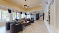 Located in Columbia, TN, this thoughtfully designed single-floor clubhouse offers a refined and welcoming atmosphere ideal for socializing and relaxing. The main lounge area is anchored by a dramatic floor-to-ceiling brick fireplace with a mounted flat-screen TV above, surrounded by plush upholstered seating arranged atop richly patterned area rugs. Tray ceilings with warm yellow accents add architectural depth, while a statement chandelier with exposed bulb rings serves as a central focal point overhead. Large windows dressed with navy blue curtains fill the space with generous natural light and offer views of the surrounding outdoors. French doors provide additional access and openness to an adjoining seating area, where curved barrel chairs and contemporary accent tables create an inviting conversation nook. The wet bar area stands out with its bold, deep-blue glossy tile backsplash, black granite countertop, and geometric pendant lights suspended above a curved bar with wood-and-metal counter stools. Light-toned geometric-patterned tile flooring flows seamlessly throughout the entire space, tying together the sophisticated navy, warm wood, and neutral color palette. Framed artwork and decorative accents complete the polished, community-focused aesthetic.