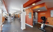 This fitness center is a comprehensively outfitted facility designed to support a full range of workout routines. The interior is defined by bold orange-painted walls accented with white structural columns and exposed beams, creating an energizing and motivating atmosphere throughout the space. Durable rubber flooring covers the entire area, providing both safety and comfort underfoot.

The cardio zone features multiple treadmills, elliptical machines, and stationary bikes positioned along mirrored walls, which help visually expand the space while allowing users to monitor their form. Bright fluorescent lighting ensures the entire facility is well-illuminated. Several windows along the perimeter allow natural light to filter in and offer views of the surrounding greenery.

The strength training area is well-stocked with a variety of cable machines, selectorized weight machines, and benches from brands including Matrix, True, and Hoist, accommodating both upper and lower body workouts. A wall-mounted television is present in the strength zone for entertainment during training sessions.

A dedicated locker alcove provides convenient secure storage. Instructional fitness charts and heart rate training guides are posted on the walls throughout, offering reference information for users. A separate entry and exit with glass-panel doors connects the facility to the outdoors, ensuring easy accessibility.