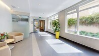 Located at 819 Yates Street in downtown Victoria, BC, this single-floor common area space comprises two interconnected hallways forming an L-shaped layout, offering a generous and welcoming environment for residents and visitors alike. The design is clean and contemporary throughout, with large-format grey and beige ceramic tiles underfoot and smooth white ceilings featuring recessed pot lighting complemented by elegant linear cove lighting along the upper walls.

The main lobby area serves as the heart of the space, featuring a built-in wood-panelled mailbox wall, a curated seating arrangement with neutral-toned lounge chairs around a central side table, and a striking piece of wall art that adds a creative focal point. Natural light pours in through large street-facing windows and glass entry doors, creating a bright and inviting atmosphere.

The secondary hallway corridor extends the space further, lined with floor-to-ceiling glossy white wall panels, additional recessed lighting, and strategically placed potted plants that introduce a refreshing natural element. Windows along one side draw in ample daylight and offer views of surrounding greenery. Vibrant yellow accent seating adds a modern, energetic touch throughout. The overall ambiance is polished, functional, and thoughtfully designed to create a positive first impression.
