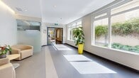 Located at 819 Yates Street in downtown Victoria, BC, this single-floor common area space comprises two interconnected hallways forming an L-shaped layout, offering a generous and welcoming environment for residents and visitors alike. The design is clean and contemporary throughout, with large-format grey and beige ceramic tiles underfoot and smooth white ceilings featuring recessed pot lighting complemented by elegant linear cove lighting along the upper walls.

The main lobby area serves as the heart of the space, featuring a built-in wood-panelled mailbox wall, a curated seating arrangement with neutral-toned lounge chairs around a central side table, and a striking piece of wall art that adds a creative focal point. Natural light pours in through large street-facing windows and glass entry doors, creating a bright and inviting atmosphere.

The secondary hallway corridor extends the space further, lined with floor-to-ceiling glossy white wall panels, additional recessed lighting, and strategically placed potted plants that introduce a refreshing natural element. Windows along one side draw in ample daylight and offer views of surrounding greenery. Vibrant yellow accent seating adds a modern, energetic touch throughout. The overall ambiance is polished, functional, and thoughtfully designed to create a positive first impression.