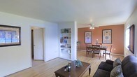 This single-floor residence offers an open-concept layout that seamlessly connects the living and dining areas, creating a cohesive and functional living space. The floors throughout are finished in light-toned hardwood, lending warmth and continuity across the rooms.

The living area features a classic wooden coffee table paired with a dark upholstered sofa, providing a comfortable and inviting gathering space. A built-in shelving unit is recessed into the dividing wall, offering practical display and storage space for books and decorative items.

The dining area is distinguished by a rich terracotta accent wall that adds depth and warmth to the space. Framed botanical artwork adorns the walls, complementing the earthy tones. A traditional dark wood dining table with upholstered chairs sits beneath a ceiling fan with light fixture, providing both comfort and illumination.

A white paneled door visible from the living area leads to an adjoining room or hallway, suggesting additional private spaces within the residence. The overall ambiance is warm, classic, and well-organized, with a balanced blend of neutral tones and warm accent colors throughout.