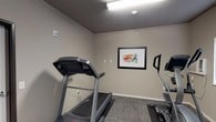 This single-floor fitness space is designed with functionality and practicality in mind, offering a dedicated area for exercise and wellness. The main workout area is equipped with a treadmill and an elliptical trainer, providing essential cardio options for residents or users. A colorful framed artwork depicting a running figure adds a motivating touch to the otherwise neutral-toned walls, giving the room a focused athletic ambiance. Overhead fluorescent lighting ensures the space is well-lit, while a smoke detector and textured ceiling contribute to a complete, safety-conscious environment. The flooring features low-pile carpet, providing a comfortable and durable surface suited to light exercise activity. A wall-mounted flat-screen television is installed in an adjacent area, ideal for entertainment during workouts or use as a lounge or media corner. The space also includes a closet area fitted with wire shelving and hanging rods, offering practical organizational storage. Window treatments with horizontal blinds provide light control and privacy. The overall design is clean and understated, with warm greige walls and white trim creating a cohesive, no-frills aesthetic that keeps the focus on utility and comfort.