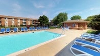 This inviting outdoor amenity space centers around a generously sized swimming pool with crystal-clear blue water, designed to accommodate both leisurely swims and relaxation. The pool area is surrounded by a wide concrete deck that provides plenty of room for movement and sunbathing. Lining one side of the pool is a row of white and blue lounge chairs, offering residents comfortable spots to unwind under the open sky. A dedicated seating area with a table and additional chairs is positioned near a brick amenity building, which appears to house pool-related facilities and posted community information. The surrounding landscape features mature trees that provide natural shade and greenery, enhancing the overall ambiance of the space. The brick construction of the adjacent buildings is consistent with the community's architectural character, lending a cohesive and well-maintained appearance throughout. The pool is clearly marked with depth indicators, reflecting attention to safety. With its open layout, abundant seating, and attractive setting, this outdoor space serves as a central social hub for the community, ideal for warm-weather recreation and relaxation.