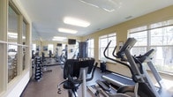 This single-level fitness center offers a thoughtfully arranged workout space designed to accommodate a range of exercise needs. The room is finished with warm, light yellow walls and a clean white ceiling fitted with recessed rectangular fluorescent lighting panels that ensure consistent, even illumination throughout the space. A large mirrored wall runs along one side of the room, visually expanding the area and allowing users to monitor their form during exercise. Multiple windows along the opposite wall allow natural light to filter in, contributing to an open and energizing atmosphere. The flooring features a durable, dark rubberized surface suited for high-traffic fitness use. Equipment includes TRUE-branded upright and recumbent cycles, treadmills, elliptical trainers, a multi-function cable machine, and a free weight rack with dumbbells. A wall-mounted flat-screen television is positioned for viewing during cardio sessions. The layout is organized to allow comfortable movement between stations, making efficient use of the available floor space. Overall, the fitness center presents a clean, functional, and well-lit environment suitable for daily use.