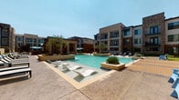 This expansive outdoor amenity space is designed to deliver a resort-caliber experience within a contemporary residential setting. At its centerpiece is a generously sized swimming pool with crystal-clear blue water, highlighted by integrated water fountain features and signature in-water chaise loungers that invite relaxation. The surrounding sun deck is finished in warm-toned paver stonework, providing ample space for lounge chairs and umbrellas positioned to capture abundant natural sunlight.

The architecture framing the space blends warm brick masonry in varied tones with modern panel siding and large-format windows, creating a visually rich and cohesive aesthetic. Raised planter beds filled with ornamental grasses and native landscaping add greenery and texture throughout the courtyard, softening the hardscape and enhancing the overall ambiance.

A welcoming entry patio — branded as "Knox Reserve" — features stylishly furnished seating with woven chairs and a metallic accent table, accessible through floor-to-ceiling glass doors and windows that connect the interior and exterior seamlessly. Glass-panel railings with dark metal frames define walkways and terraced transitions between zones, maintaining open sightlines across the courtyard. The space is bathed in natural light and designed for both leisurely lounging and vibrant social gatherings.