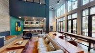 This thoughtfully designed community amenity space combines sophisticated aesthetics with a versatile, social atmosphere across a single open floor. Soaring double-height ceilings define the main lounge area, where an expansive wall of floor-to-ceiling glass doors floods the interior with natural light and provides seamless access to a landscaped outdoor courtyard. The open layout accommodates distinct activity zones, including a relaxed lounge area outfitted with a plush sectional sofa, a patterned area rug, and a low-profile coffee table. Adjacent to the lounge, a shuffleboard table and classic arcade cabinet offer recreational entertainment, while a pinball machine adds a playful retro touch near the entry. The kitchenette area is a design highlight, featuring a bold teal accent wall, a large-format abstract artwork, open brass shelving with white tile backsplash, stainless steel appliances, and a generous island with woven leather barstools. A separate lounge nook showcases warm horizontal wood-paneled walls, navy blue cabinetry, built-in banquette seating in cognac leather, a marble-topped side table, and a wall-mounted flat-screen TV. Warm hardwood flooring, bold pendant lighting, and carefully curated artwork tie the space together into a cohesive, design-forward environment.