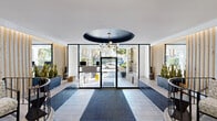 This thoughtfully designed lobby combines contemporary and mid-century modern influences to create a welcoming and visually dynamic entry space. At its center, a bold navy oval ceiling recess anchors the room, from which a multi-arm splay chandelier with globe lights descends, serving as a key focal point. Floor-to-ceiling black-framed glass panels frame the main entrance, flooding the space with natural light and offering clear views of the surrounding landscaping and streetscape beyond.

The floor is finished in large-format light stone tiles, providing a clean and neutral base that complements the layered design elements throughout. Flanking the entrance are raised platform areas clad in deep navy glazed brick tiles, edged with curved wrought iron railings and adorned with tall snake plants in matte black planters. Vertical natural wood slat paneling lines the side walls, introducing warmth and texture that balances the sleekness of the tiled surfaces.

Toward the rear of the lobby, a seating area transitions into an elevator vestibule, where soft teal walls are decorated with striking gold and silver sunburst wall sculptures. A front desk area is visible near the entrance, completing this polished, functional, and design-forward reception space.