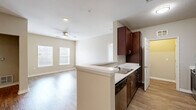 This single-floor, 2-bedroom, 1-bathroom unit offers a thoughtfully designed layout that makes efficient use of its space. The open-concept kitchen and living area serves as the heart of the home, where rich dark-wood cabinetry with brushed nickel hardware contrasts beautifully against light countertops and a peninsula with a double sink. Stainless steel appliances, including a refrigerator and dishwasher, add a modern touch. Wood-style flooring runs throughout the main living and kitchen areas, while two large windows allow natural light to fill the living room, which is also equipped with a ceiling fan. A dedicated hallway connects the living spaces to the private quarters. The primary bedroom is a generous room featuring plush carpeting, a ceiling fan, and a window with natural light. It is complemented by a walk-in closet outfitted with wire shelving and hanging rods for ample storage. The second bedroom also offers carpeted flooring and a window. The bathroom features dark wood cabinetry with a vanity and well-lit mirror. Throughout the unit, four closets provide exceptional storage capacity. The neutral warm-beige color palette and clean, consistent finishes create a cohesive and comfortable living environment.
