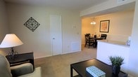 This single-floor residence offers a practical and comfortable open-concept layout, with the living and dining areas flowing seamlessly into one another. The living room features wall-to-wall carpeting in a warm neutral tone, providing a soft and inviting foundation for the space. A decorative wrought-iron wall accent adds a touch of character to the otherwise clean, understated walls painted in a soft off-white. A white interior door provides access to an additional room, while a floor-level vent indicates a central HVAC system throughout. The adjacent dining area is defined by a round table set with dark wood chairs and is anchored overhead by a classic chandelier with warm-toned bulbs, contributing an intimate ambiance. A framed piece of wall art adds a decorative touch to the dining space. The transition between the two areas is marked by a subtle architectural beam and a partial white countertop or divider, suggesting proximity to a kitchen. Overall, the space presents a cohesive, neutral palette that is well-suited for comfortable everyday living.