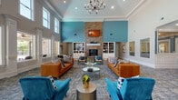 This single-floor community clubhouse is designed with an open, welcoming layout that blends elegance with comfort. The grand entrance foyer greets visitors with light hardwood-style flooring, soaring double-height ceilings, white decorative columns, and a statement chandelier set against a teal-accented ceiling inset — establishing a refined first impression.

The expansive main lounge is the centerpiece of the space, featuring dramatic ceiling heights, a floor-to-ceiling stacked stone fireplace with a white mantel and wall-mounted television, and flanking built-in shelving units. Teal accent walls complement richly upholstered cognac leather sofas and vibrant teal velvet armchairs, creating a bold yet inviting atmosphere. Patterned area rugs anchor multiple seating arrangements throughout.

A wall of tall, column-framed windows floods the lounge with natural light and provides direct views of the outdoor pool area, with access via French doors. Additional seating nooks offer more intimate settings, including a cozy lounge corner with teal barrel chairs, geometric mirrors, and a dedicated coffee station. A separate dining area features a round table beneath an amber glass chandelier, surrounded by lush outdoor views. The space is thoroughly detailed with crown molding, recessed lighting, and curated artwork throughout.