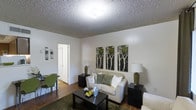 This single-floor residence offers a comfortable and thoughtfully arranged living space designed for everyday functionality and ease. The living room serves as the heart of the home, featuring warm hardwood flooring, a textured ceiling, and neutral white walls that create a bright, clean backdrop. A plush sofa accented with decorative pillows anchors the seating area, complemented by a dark wood coffee table and coordinating end tables with table lamps that provide warm ambient lighting. A set of nature-inspired wall art panels depicting birch trees adds a subtle organic touch to the room's décor. A green area rug grounds the space and adds a pop of color, while a small dining area with green chairs is tucked neatly to one side, making efficient use of the available floor space. A doorway leads to additional rooms, suggesting a practical and connected floor plan. The overall ambiance is cozy and welcoming, with a classic residential aesthetic that balances comfort and style. The space is well-lit through a central ceiling fixture, contributing to an open and airy feel throughout.