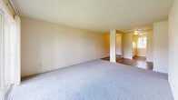 This single-floor unit in Vancouver, Washington, features a practical and efficient layout accommodating three bedrooms, one full bathroom, and a connected living and kitchen area. The living room is the largest shared space, finished with neutral-toned carpet, a textured ceiling, and a sliding glass door that opens to a private balcony with an outdoor railing — allowing natural light and exterior views to enhance the room's open feel. An adjacent area provides a transition into the kitchen, which includes light wood cabinetry and standard appliances. Each of the three bedrooms is carpeted and finished with neutral walls and textured ceilings. Two bedrooms feature sliding double-door closets, while the primary bedroom includes a window with natural light, a baseboard heater, and an electrical panel. Additional bedrooms also feature windows with outdoor views and baseboard heating. The full bathroom is equipped with a tub-shower combination, a toilet, tile flooring, and a vanity with a granite-style countertop and wood cabinetry. A central hallway connects the bedrooms to the main living areas, completing the straightforward and functional floor plan.
