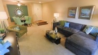 This single-floor space combines a living area and dining space in an open, connected layout. The living area is anchored by a large dark gray sectional sofa accented with blue and green throw pillows, paired with a sleek glass-top coffee table resting on a dark base. A framed piece of artwork hangs on the wall above the sofa, and a contemporary table lamp provides warm ambient lighting. The neutral beige carpeting runs throughout, contributing to a cozy and cohesive atmosphere. A side table with a lamp and small décor items sits adjacent to the sectional.

The dining area, visible to the left, features a round table surrounded by green upholstered chairs, adding a pop of color to the space. A small Christmas tree decorates the corner, adding a seasonal touch. A raised entryway with wooden steps and a white door is centrally positioned, offering a defined transition point into the space. The walls are painted in soft, warm neutral tones, keeping the overall ambiance light and inviting. The layout makes efficient use of the available space, offering distinct zones for lounging and dining within a single open floor plan.