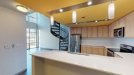 This two-floor space showcases a thoughtfully designed interior that blends modern industrial aesthetics with functional living areas. At the heart of the main level is a well-appointed kitchen featuring light maple cabinetry, crisp white countertops, and a full suite of stainless steel appliances including a range, built-in microwave, and refrigerator. A bold yellow ceiling energizes the kitchen area, complemented by pendant lighting and recessed track lights that illuminate the space effectively. A generous kitchen island with an integrated sink provides ample workspace and serves as a natural dividing element.

A dramatic spiral staircase with a sleek metal railing serves as both a functional and sculptural centerpiece, connecting the two floors with architectural flair. The lower level features soaring ceilings with exposed structural decking, large windows that invite abundant natural light, and views to the outdoors. The upper floor opens onto a landing with patterned carpet flooring, multiple doorways, and continues the industrial-modern aesthetic with exposed ceiling elements. Throughout the space, a neutral color palette of whites and grays is punctuated by bold accent colors, creating a contemporary and visually engaging environment.