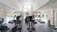 Located at 6312 Reafield Dr in Charlotte, NC, this fitness center is housed in a single-story facility that combines thoughtful architectural design with a comprehensive array of exercise equipment. The space is defined by its dramatic vaulted ceiling with a striking floor-to-ceiling triangular window, flooding the cardio area with abundant natural light and offering views of an outdoor pool and surrounding community. White shiplap-style ceiling panels and crisp trim work lend a clean, modern aesthetic throughout.

The cardio zone is well-appointed with multiple treadmills, elliptical trainers, and stair climbers arranged to take full advantage of the soaring windows and natural light. An adjacent strength and functional training area offers a multi-station cable machine, a full rack of dumbbells, adjustable weight benches, and a diverse selection of functional training tools including stability balls, medicine balls, and a target training station. Durable rubber flooring spans the workout areas, providing both safety and comfort.

Large French doors open to an outdoor patio and lounge area, seamlessly connecting the interior to the outdoors. Recessed and track lighting ensures the space is well-lit at all hours. The overall ambiance is bright, airy, and motivating, making this fitness center a standout amenity for the Reafield Drive community.